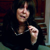A woman with long dark hair leans on a green surface, holding eyeglasses near her mouth and gazing thoughtfully. She wears jewelry inspired by crystal magick and has The Witch's Oracle, 2nd Edition by Schiffer Publishing, Ltd. beside her.