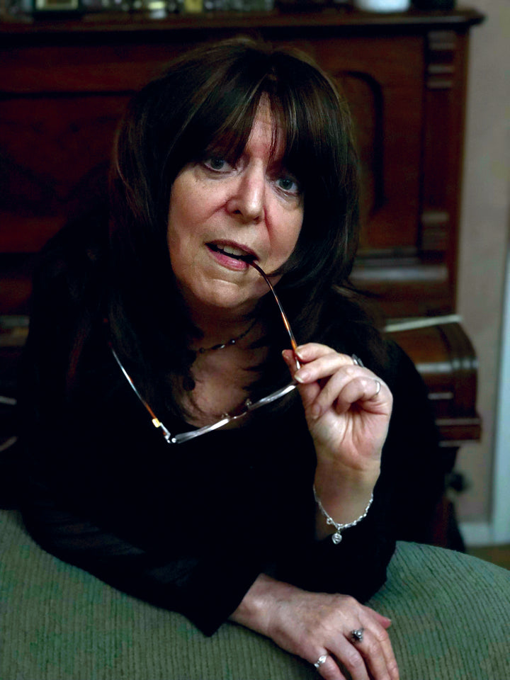 A woman with long dark hair leans on a green surface, holding eyeglasses near her mouth and gazing thoughtfully. She wears jewelry inspired by crystal magick and has The Witch's Oracle, 2nd Edition by Schiffer Publishing, Ltd. beside her.