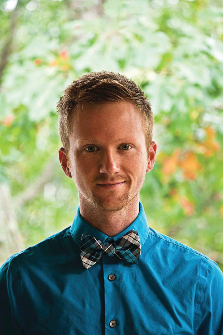A man with short light brown hair in a bright blue shirt and plaid bow tie stands outside with greenery, smiling slightly while holding The Witch’s Oracle, 2nd Edition by Schiffer Publishing, Ltd.