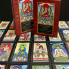 The Buddha Tarot by Schiffer Publishing, Ltd. features vibrant cards with Major Arcana symbolism spread on a surface, while its red box and guidebook with an illustrated Buddha stand upright at the center, evoking enlightenment.