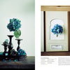 A blue hydrangea in a vase sits on dark candlesticks with petals and rocks to the left, while a single pressed hydrangea lies in an open book—a perfect scene inspired by The Herbarium of Fabric Flowers from Schiffer Publishing, Ltd.
