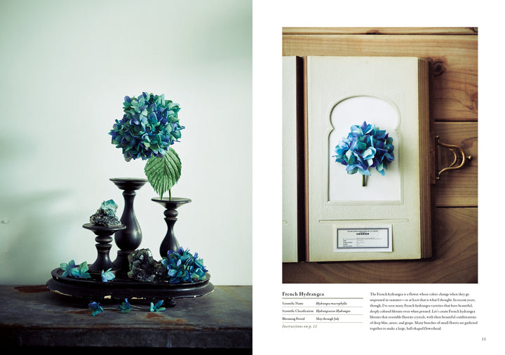 A blue hydrangea in a vase sits on dark candlesticks with petals and rocks to the left, while a single pressed hydrangea lies in an open book—a perfect scene inspired by The Herbarium of Fabric Flowers from Schiffer Publishing, Ltd.