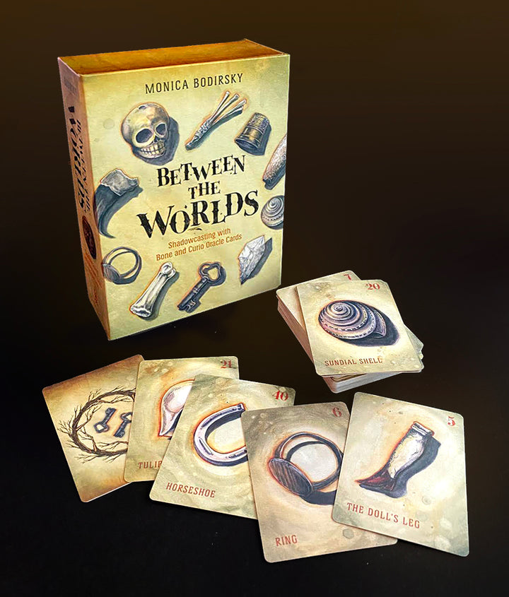 A box labeled Between the Worlds by Schiffer Publishing, Ltd. stands upright on a dark surface, surrounded by illustrated oracle cards depicting objects such as a ring, doll’s leg, horseshoe, sundial shell, and wishbone.