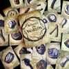 A deck of illustrated divination cards, "Between the Worlds" by Schiffer Publishing, Ltd., features mystical items like a skull, raven, doll’s head, and bones surrounding a central card.
