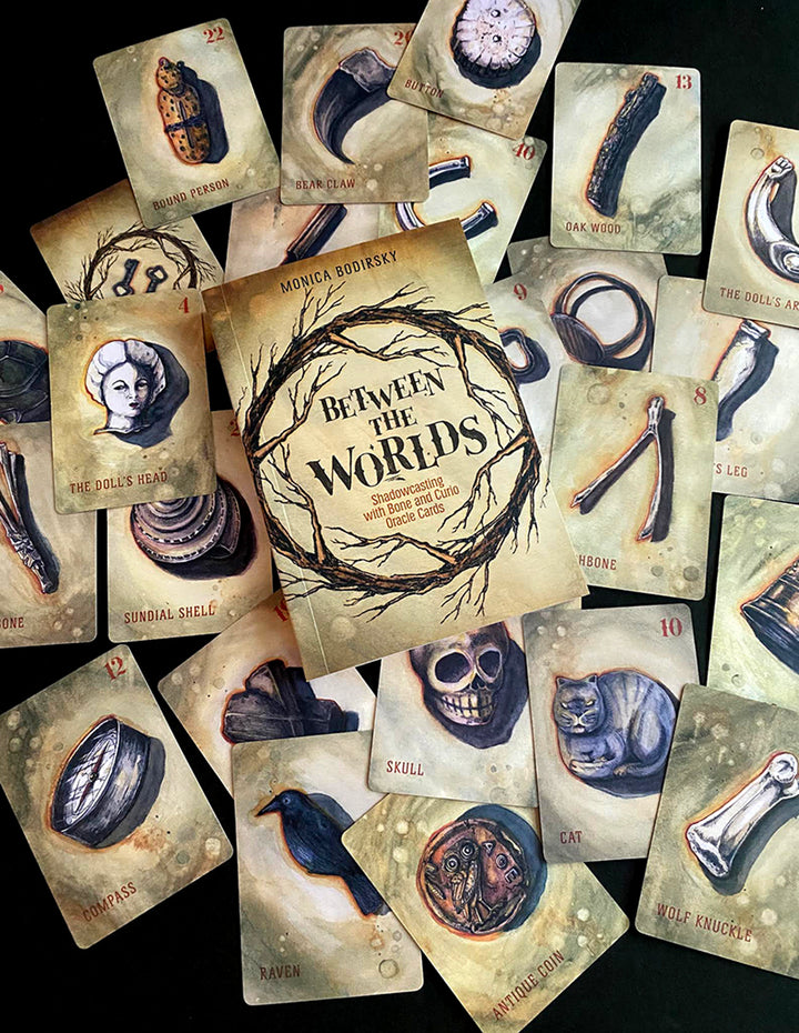 A deck of illustrated divination cards, "Between the Worlds" by Schiffer Publishing, Ltd., features mystical items like a skull, raven, doll’s head, and bones surrounding a central card.