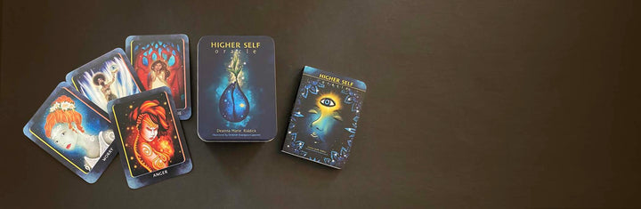 Several illustrated spiritual guidance cards from the Higher Self Oracle by Schiffer Publishing, Ltd. are spread out with their box on a dark surface, featuring designs like a blue teardrop and an eye.