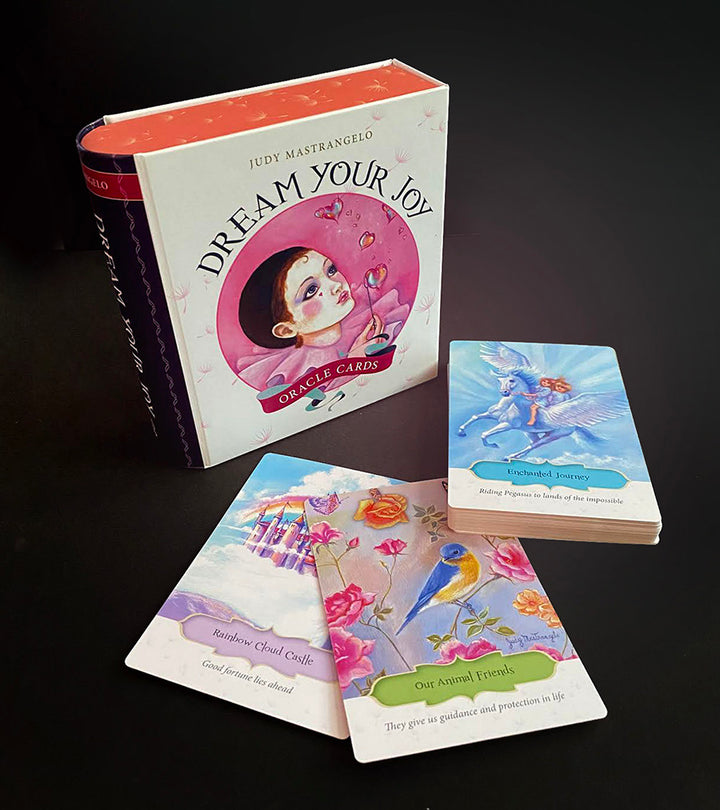 A box of Dream Your Joy Oracle Cards by Schiffer Publishing, Ltd. sits beside three colorful affirmation cards with whimsical, Mind Painting-inspired illustrations and positive messages, all arranged on a black background.
