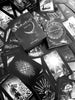 A black and white image displays the Nightfall Tarot by Schiffer Publishing, Ltd., featuring its deck box surrounded by all 78 uniquely illustrated cards arranged in a scattered Rider-Waite-Smith-inspired pattern.