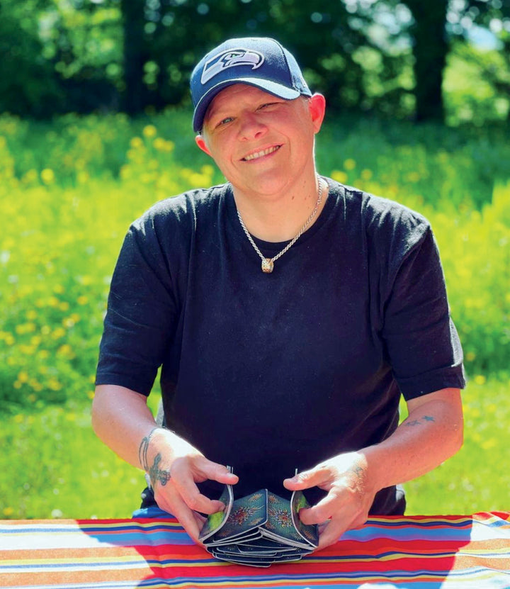 A person in a navy Seattle Seahawks cap and black shirt sits at a striped outdoor table, smiling as they shuffle Sigils tarot cards by Schiffer Publishing, Ltd., with greenery and yellow flowers in the background.
