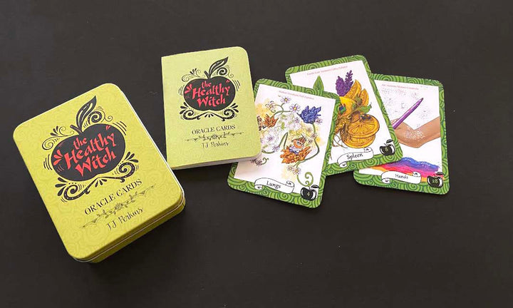 The Healthy Witch Oracle Cards by Schiffer Publishing, Ltd. are shown on a black surface with the box, guidebook, and three cards—Lemon, Squash, and Rainbow—each highlighting unique magick correspondences.