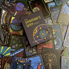 A deck of vibrant ascension cards with mystical hand-drawn art is displayed, featuring The Shaman’s Way Oracle by Schiffer Publishing, Ltd. at the center—an ideal tool for spiritual healing.