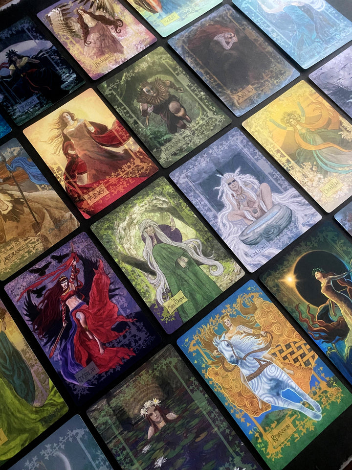 A spread of the Celtic Goddesses, Witches, and Queens Oracle by Schiffer Publishing, Ltd., features vibrant cards with mythological Celtic figures artfully arranged diagonally on a dark surface as a striking divination tool.