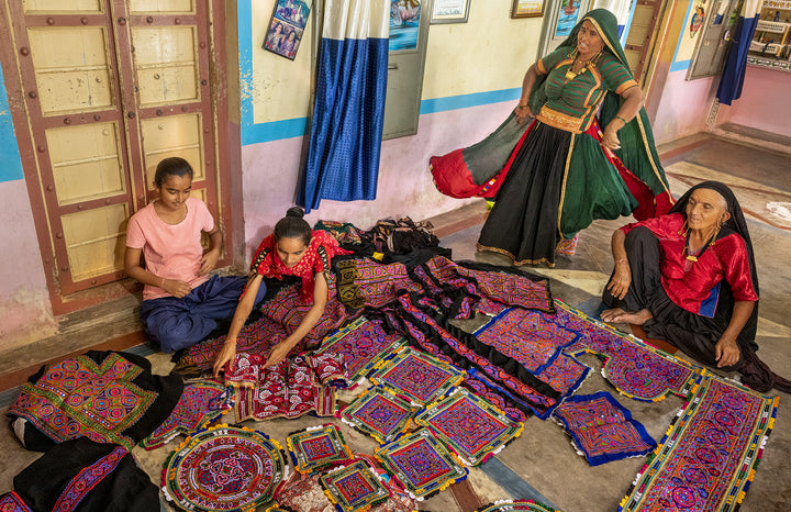 Four women in traditional attire, representing Indian artisans, craft and converse among vibrant embroidered textiles. Artisans by Design by Schiffer Publishing, Ltd. captures their creativity in a room full of colorful handmade beauty.