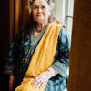 An older woman with gray hair in two buns, wearing a patterned teal dress and yellow scarf, sits by a window smiling gently, holding The Magic Lantern Sibilla oracle cards by Schiffer Publishing, Ltd., bathed in natural light.