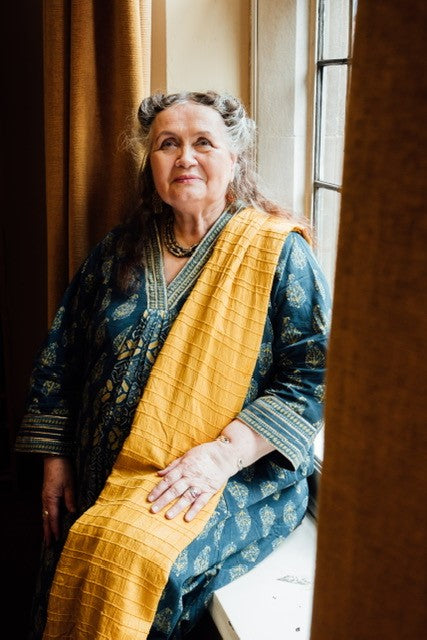 An older woman with gray hair in two buns, wearing a patterned teal dress and yellow scarf, sits by a window smiling gently, holding The Magic Lantern Sibilla oracle cards by Schiffer Publishing, Ltd., bathed in natural light.