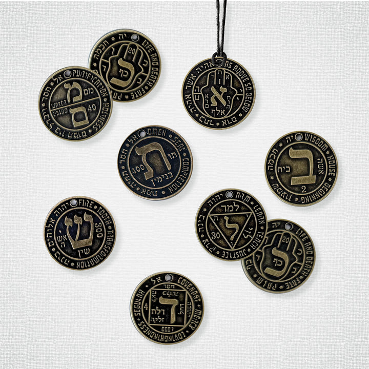 Nine bronze Hebrew Casting Lots by Schiffer Publishing, Ltd., engraved with the Hebrew Alefbet and English words like Wisdom, Justice, and Omen, are displayed on a white surface. One is shown on a black cord.