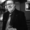 A man with glasses, beard, and mustache stands indoors in a suit and tie, holding a wine glass. The black-and-white image evokes the mysterious atmosphere of The John Dee Oracle from Schiffer Publishing, Ltd.