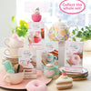 A display of Schiffer Publishing, Ltd.’s Crochet Cafe Amigurumi Kit Hot Cocoa with cupcakes, donuts, pastel yarn, stacked teacups, and a floral teapot. Text: “Collect the whole set!”.