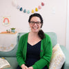 A woman with dark curly hair and glasses smiles on a light blue couch, with pastel felt wall decor behind her. Featured: "HeartFELT Crafts to Make and Give" by Schiffer Publishing, Ltd., inspiration for handmade home accents.