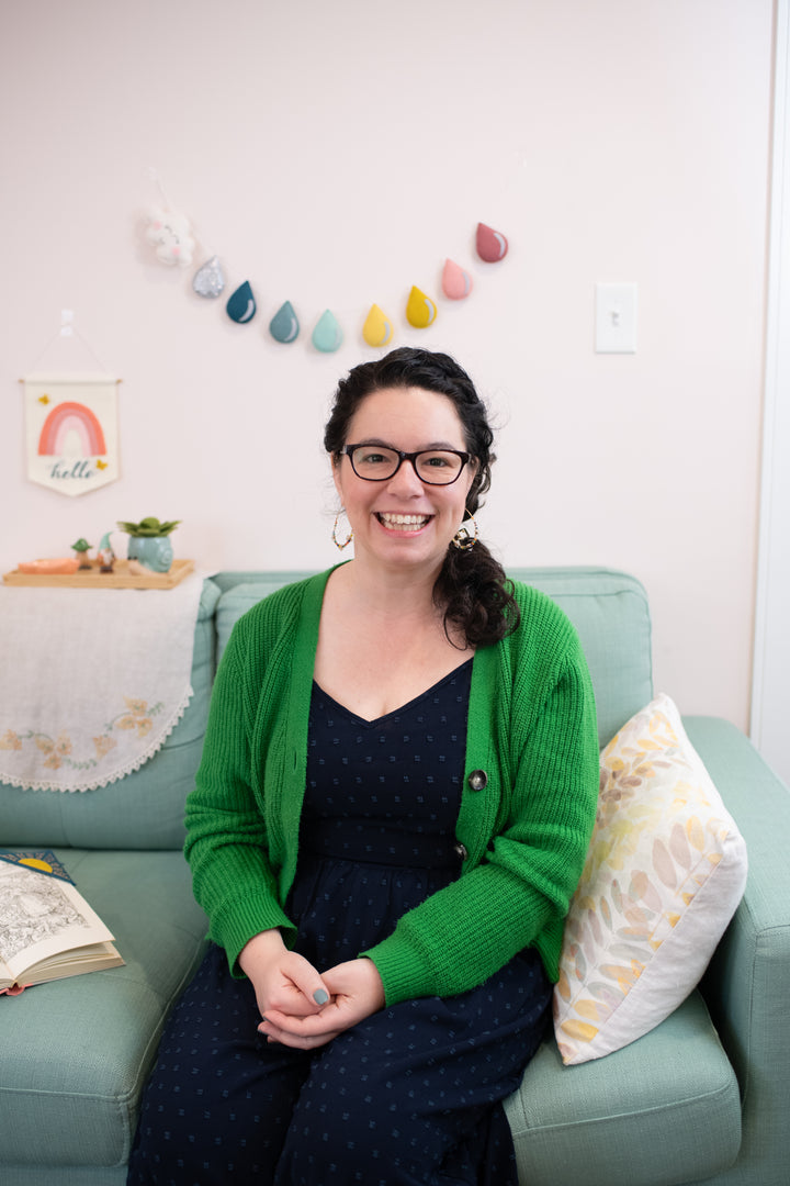 A woman with dark curly hair and glasses smiles on a light blue couch, with pastel felt wall decor behind her. Featured: "HeartFELT Crafts to Make and Give" by Schiffer Publishing, Ltd., inspiration for handmade home accents.