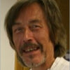 A middle-aged man with shoulder-length brown hair, a mustache, and a beard smiles in a white shirt. The softly blurred background evokes the timeless mysteries found in The Camelot Oracle by Schiffer Publishing, Ltd.