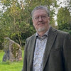 An older man with glasses, gray beard, and mustache stands outdoors in a brown blazer and patterned shirt. Green grass, trees, and a mossy rock evoke the mystique of Arthurian legends featured in The Camelot Oracle by Schiffer Publishing, Ltd.