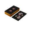A stack of Nocturnal Garden Lenormand cards by Schiffer Publishing, Ltd. is shown with gold edges. One card displays a teapot, cup, and heart-shaped steam in whimsical art; the card backs have celestial and floral designs on black.