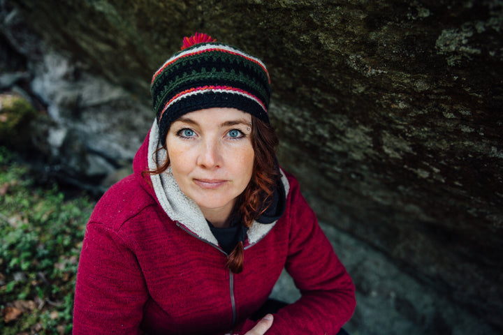 A woman with blue eyes and red braided hair sits outdoors by a large rock, wearing a red jacket and knit hat, appearing immersed in Nordic mystery as she uses the Nordic Forest Rituals Oracle Cards from Schiffer Publishing, Ltd.