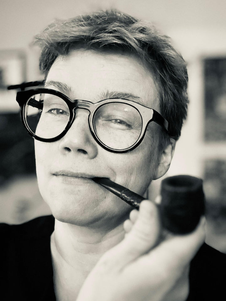A person with short hair and round glasses smiles slightly while holding a pipe, evoking the quiet mystery of forest spirits—an atmosphere captured by Schiffer Publishing, Ltd.'s Nordic Forest Rituals Oracle Cards.
