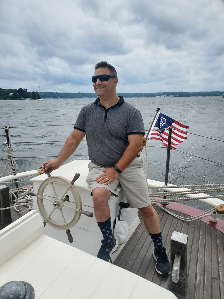 A man in sunglasses and casual attire steers a boat with an American flag on a cloudy day, capturing the spirit found in Schiffer Publishing, Ltd.'s "American Merchant Seaman's Manual, 8th Edition.