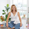 A smiling blonde woman sits cross-legged on a wooden table in a bright room with art supplies, flowers, and plants. She wears a white top, blue jeans, colorful necklaces—and has the Schiffer Publishing Watercolor the Easy Way Paint-Your-Own Watercolor Cards Garden kit nearby.