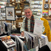 A smiling man in a striped sweater and scarf signs his book, "New Orleans" by Schiffer Publishing, Ltd., at a bookstore table surrounded by open books, Mardi Gras displays, gift items, and shelves capturing the French Quarter’s spirit.