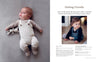 A baby in Nordic-inspired knitwear from "Baby Knits from Norway" by Schiffer Publishing, Ltd. lies on a soft gray surface with a stuffed bunny, while a toddler in dark overalls and sweater sits nearby, looking off to the side.