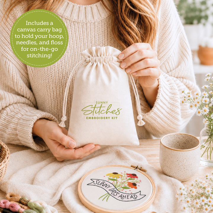 A woman holds a drawstring canvas bag labeled “Sunny Stitches Embroidery Kit Sunny Days Ahead” from Schiffer Publishing, Ltd. at a table with an embroidery hoop, needle, floss, and a finished “Sunny Days Ahead” floral embroidery.