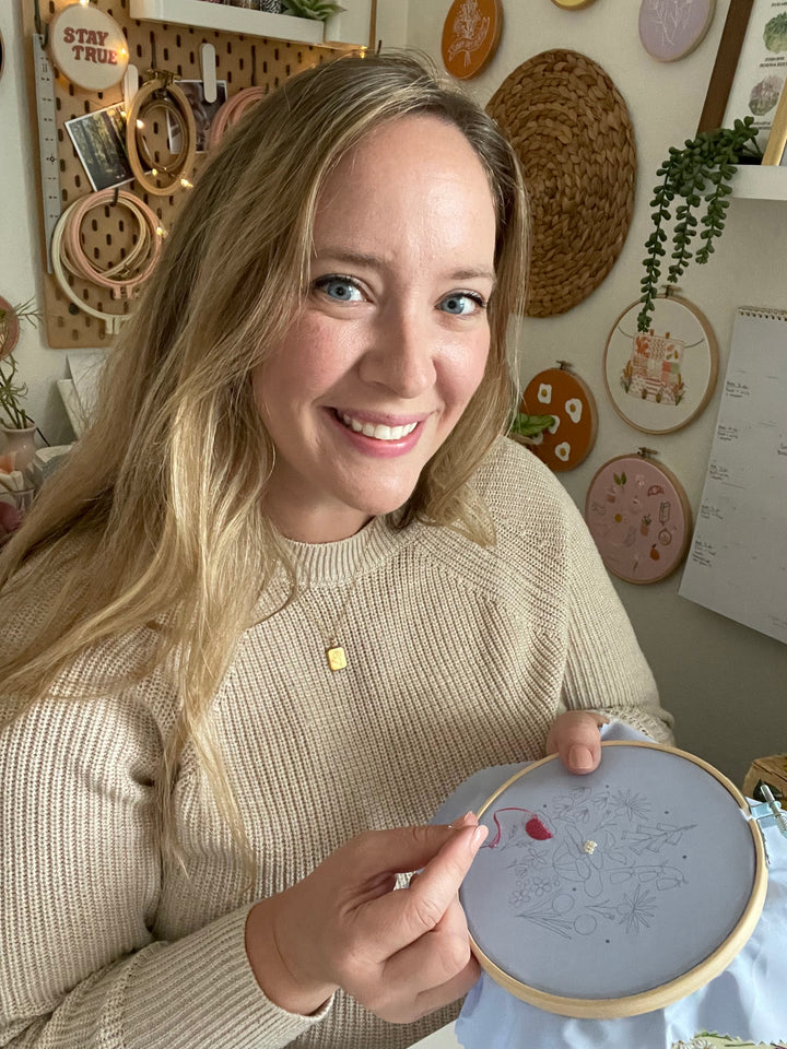 A smiling woman with long blonde hair holds the Sunny Stitches Embroidery Kit Sunshine Pines by Schiffer Publishing, Ltd., working on colorful stitches in a cozy craft room decorated with nature embroidery art on the wall behind her.
