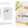 The Sunny Stitches Embroidery Kit Sunshine Pines from Schiffer Publishing, Ltd. displays a hand-embroidered landscape with pines, a river, sun, and clouds. A woven basket and green plant are nearby; supplies and instructions are shown on the left.