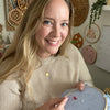 A woman with long blonde hair in a beige sweater smiles while working with a needle on the Sunny Stitches Embroidery Kit Woodland Mushroom by Schiffer Publishing, Ltd. in a cozy craft room decorated with embroidery hoops.
