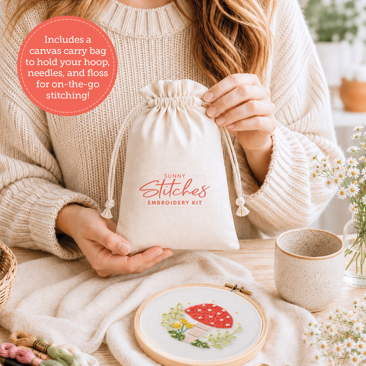 A person in a cream sweater holds the Schiffer Publishing, Ltd. Sunny Stitches Embroidery Kit Woodland Mushroom bag at a table with an embroidery hoop, threads, and a cup. The red label reads: Includes canvas carry bag for on-the-go projects!.