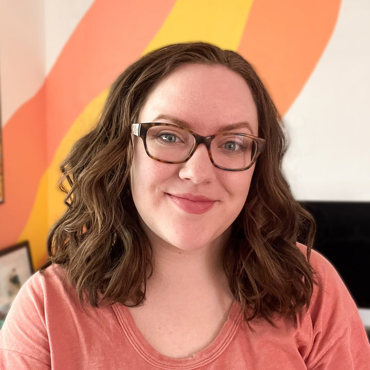 A woman with wavy brown hair and glasses smiles at the camera, wearing a peach shirt. Behind her, bold orange and yellow stripes brighten the white wall, capturing the playful spirit of a child’s painting as seen in the Schiffer Publishing, Ltd. I Heart Art Forest Friends Watercolor Paint Pad.