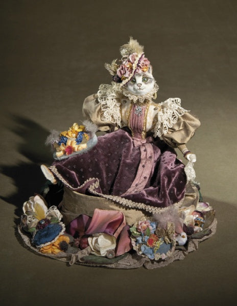 Whimsical Elegance by Schiffer Publishing, Ltd. is a handcrafted cat doll dressed in a Victorian-style gown with lace, velvet, and floral details, surrounded by vibrant fabric flowers atop a brown surface.