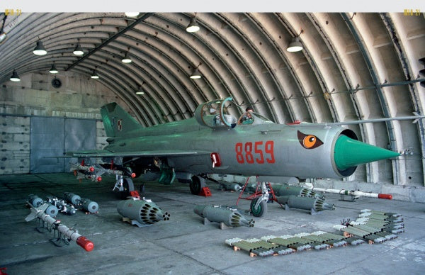 The MiG-21 by Schiffer Publishing, Ltd. features a silver and green supersonic jet numbered 8859 with a painted nose eye, displayed in an arched hangar amid bombs, missiles, and ammunition.