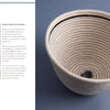 A hand displays an Artisan Ropework basket by Schiffer Publishing, Ltd., featuring black thread details and a knotted end. Beside it, a finished cream cotton rope basket with a black rim rests on a gray background.