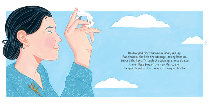 A woman with dark hair holds a bone aloft, gazing through its hole as clouds pass above. “A Bone for Bo” by Schiffer Publishing, Ltd. tells how Bo, her dog, drops the bone in her lap under the inspiring New Mexico sky.
